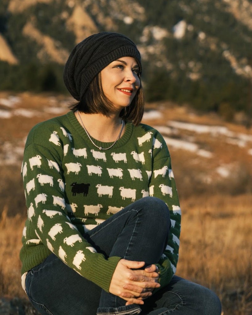 Jade Proudman sitting outdoors in front of mountains, wearing a beanie and a green jumper covered in white sheep with one black sheep